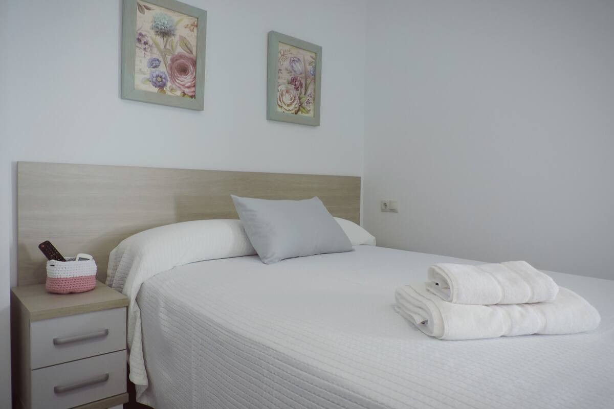 A neatly made bed with a white comforter and pillows, a bedside table with a pink basket, and two floral artwork pieces on the wall.