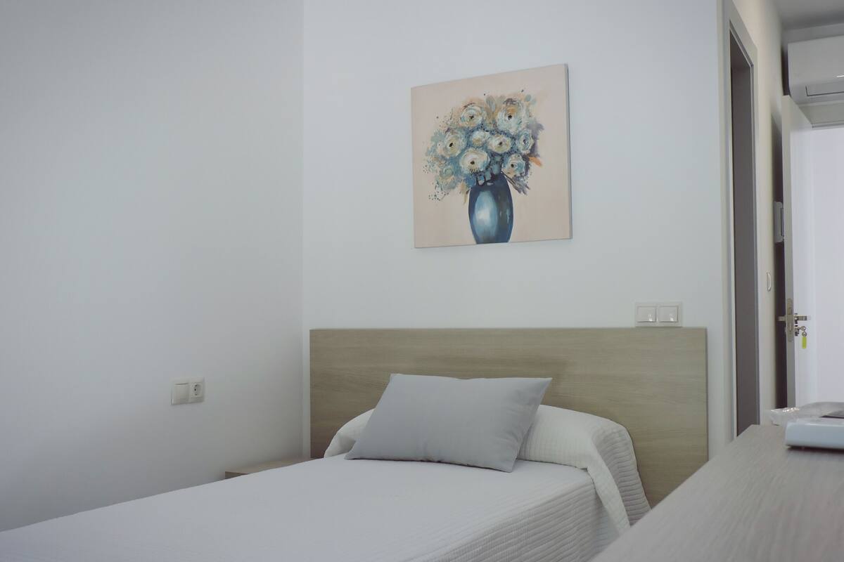A bedroom with a bed, a nightstand, a painting of flowers on the wall, and a door to the right.