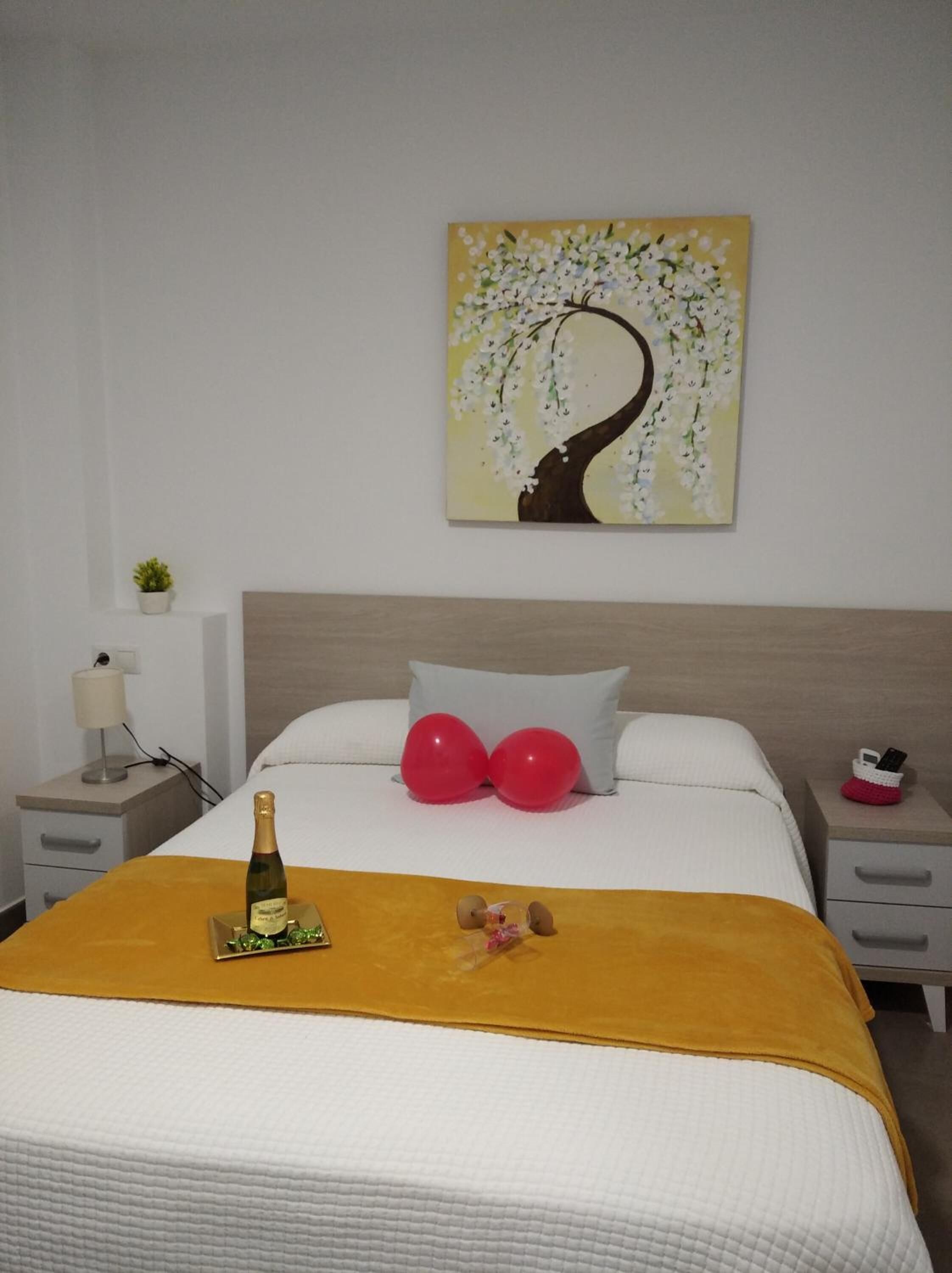 Una habitación de hotel con una cama, una mesita de noche con lámpara, una planta pequeña en maceta, un cuadro de un árbol con flores y una botella de champán sobre la cama.