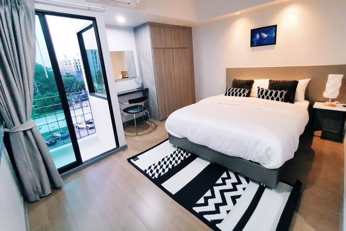 A modern hotel bedroom with a large bed, a desk, and a view of the city through a sliding door.