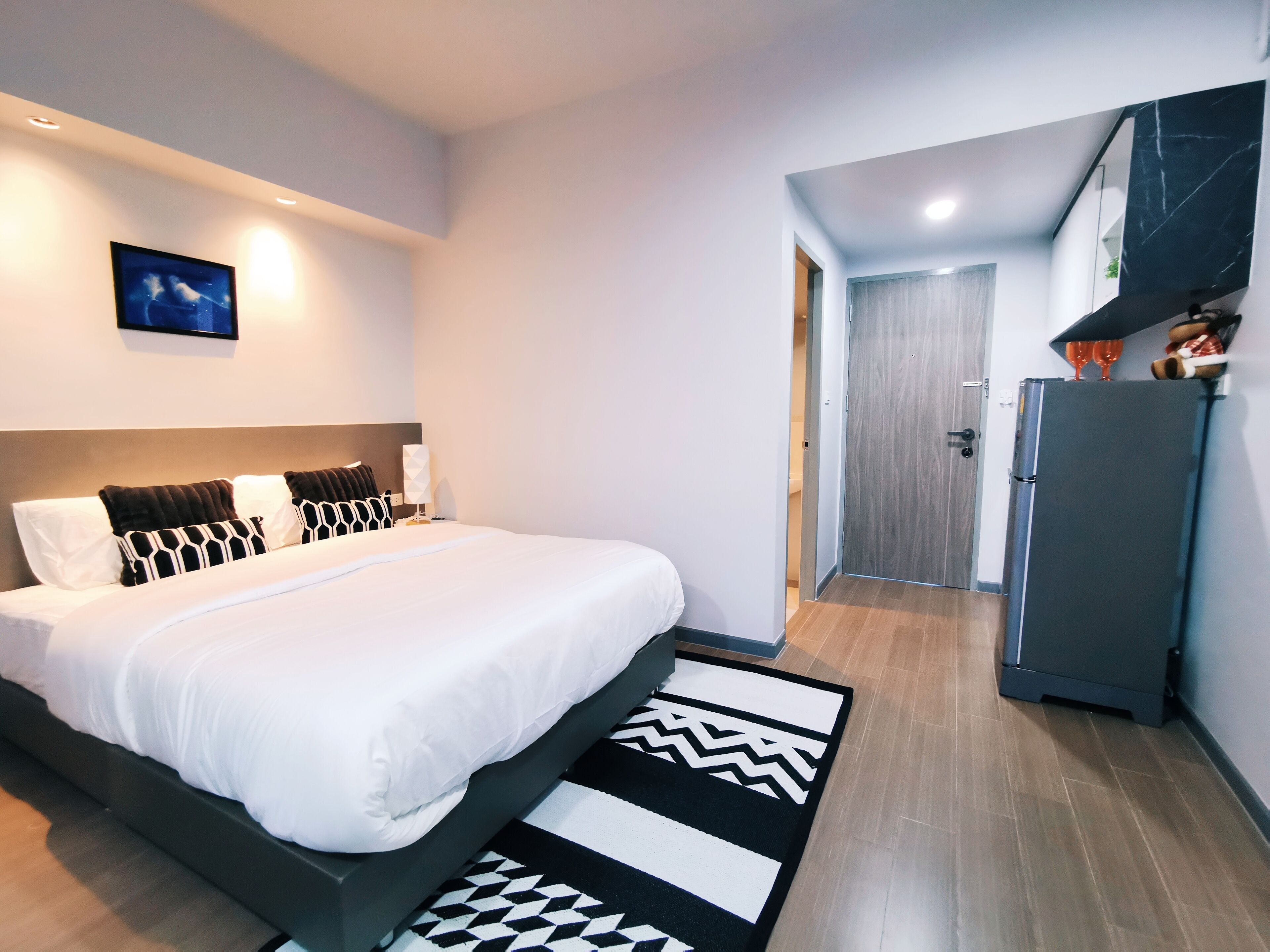 Una habitación de hotel moderna con una cama grande, un televisor de pantalla plana montado en la pared, una puerta de madera y un refrigerador.