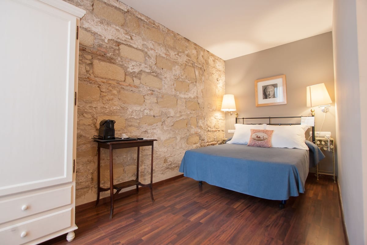 Une chambre avec un mur en pierre, un lit recouvert d’une courtepointe bleue, deux lampes de chevet, une table de chevet et une commode.