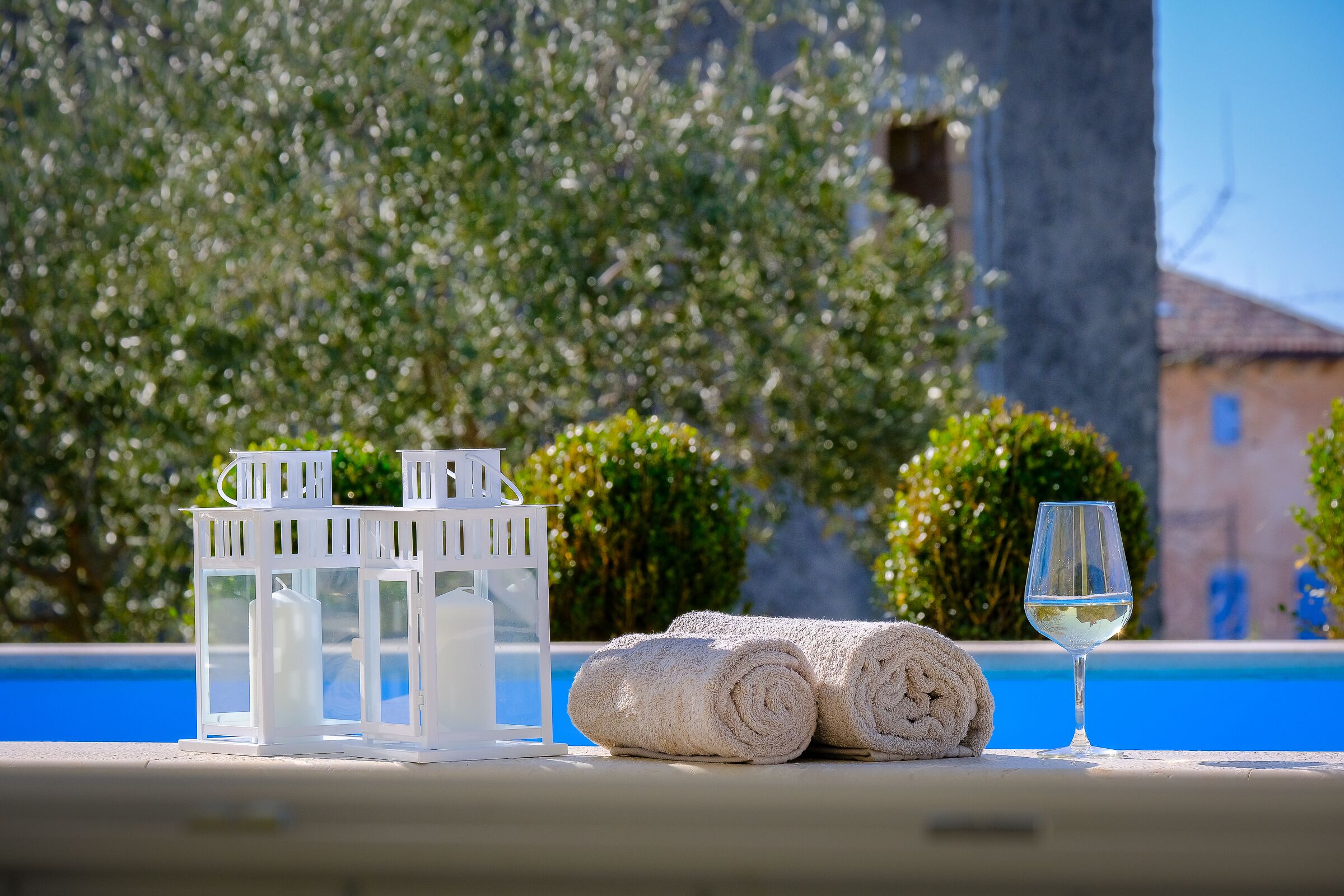 A poolside setting with a white lantern, rolled towels, and a glass of white wine.