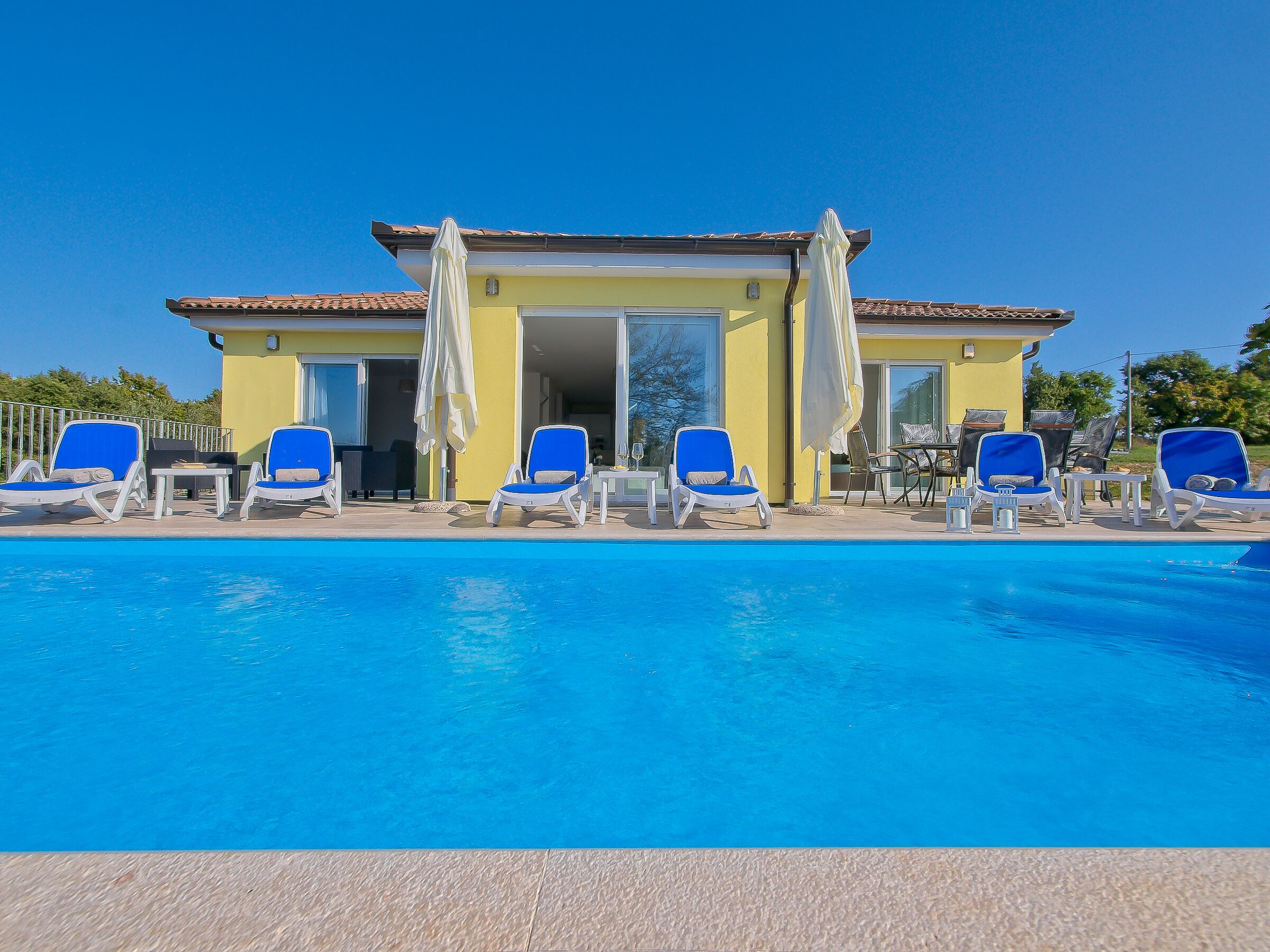 Ein Poolbereich mit blauen Liegestühlen und Sonnenschirmen, ein gelbes Gebäude mit einem Ziegeldach und einem wolkenlosen, blauen Himmel.