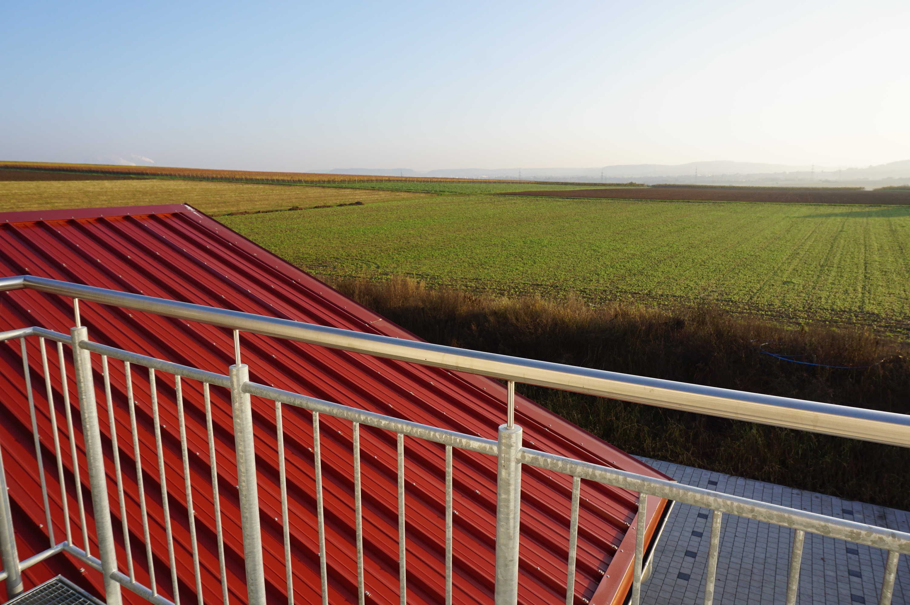 A red-roofed building with a metal railing, overlooking a vast green field.