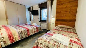 A room with two beds, each with a quilted bedspread, a television mounted on the wall, and a window with curtains. - Villa Linda - ONE-BEDROOM APARTMENT sleeps 5 (Bardonecchia)