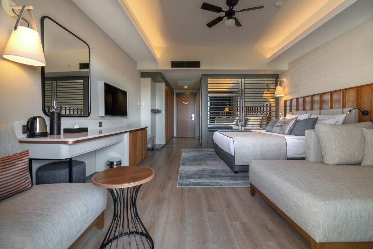A modern hotel room with a bed, a desk, a chair, a small table, and a ceiling fan.