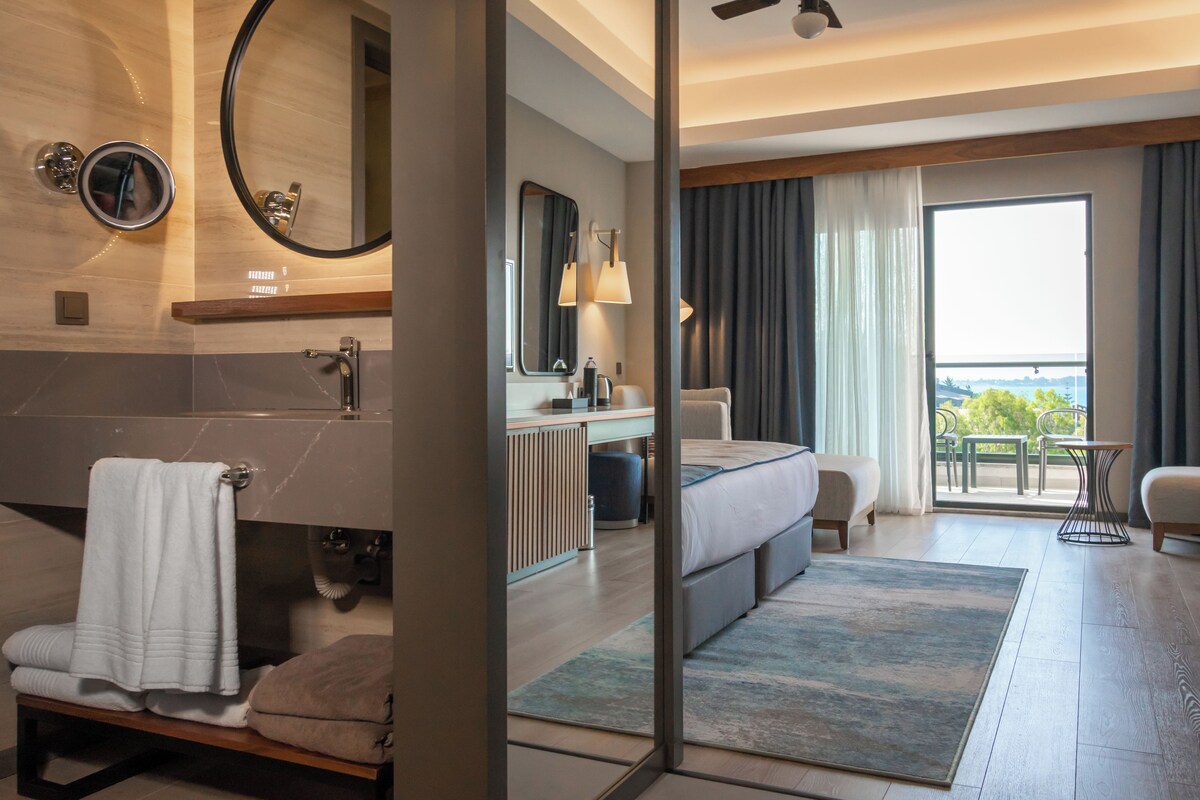 A modern hotel room with a bathroom, a bed, a sitting area, and a balcony with a view.