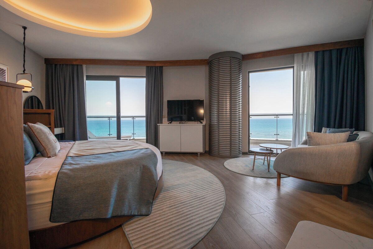 A modern hotel room with a bed, a TV, a seating area, and a view of the sea.