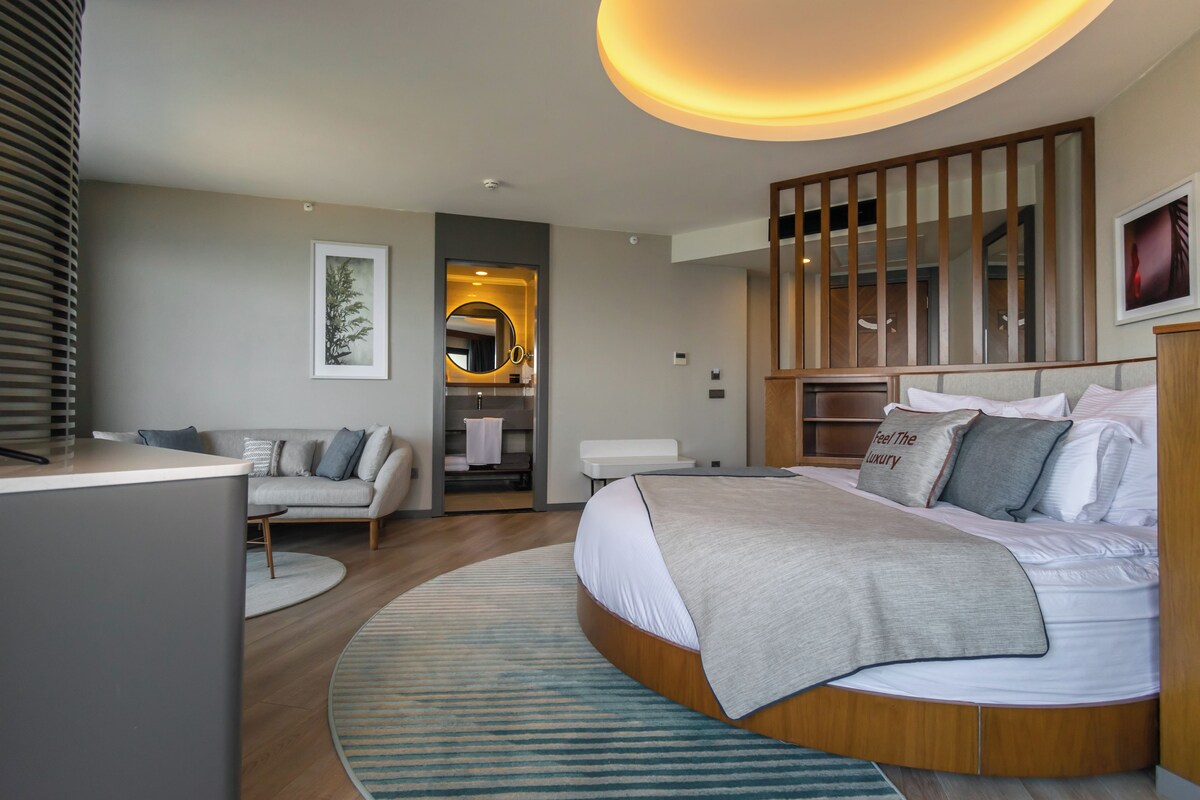 A modern bedroom with a circular bed, a sofa, a desk, and a bathroom with a mirror and towel rack.