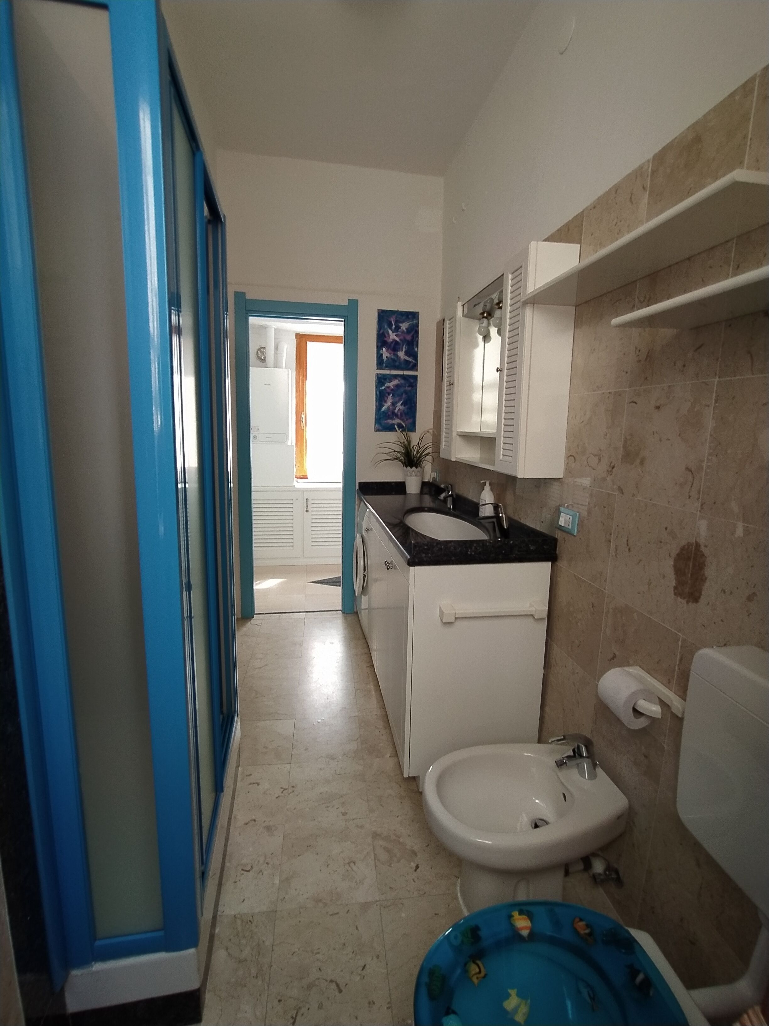 Un bagno con lavandino, servizi igienici e zona doccia. Si nota una porta blu sulla sinistra e un mobile bianco sopra il lavandino.