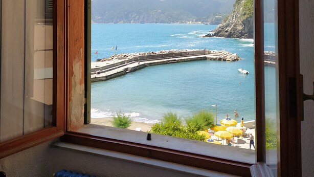 A room with a large window overlooking a coastal area, featuring a view of the sea, a pier, and distant mountains.