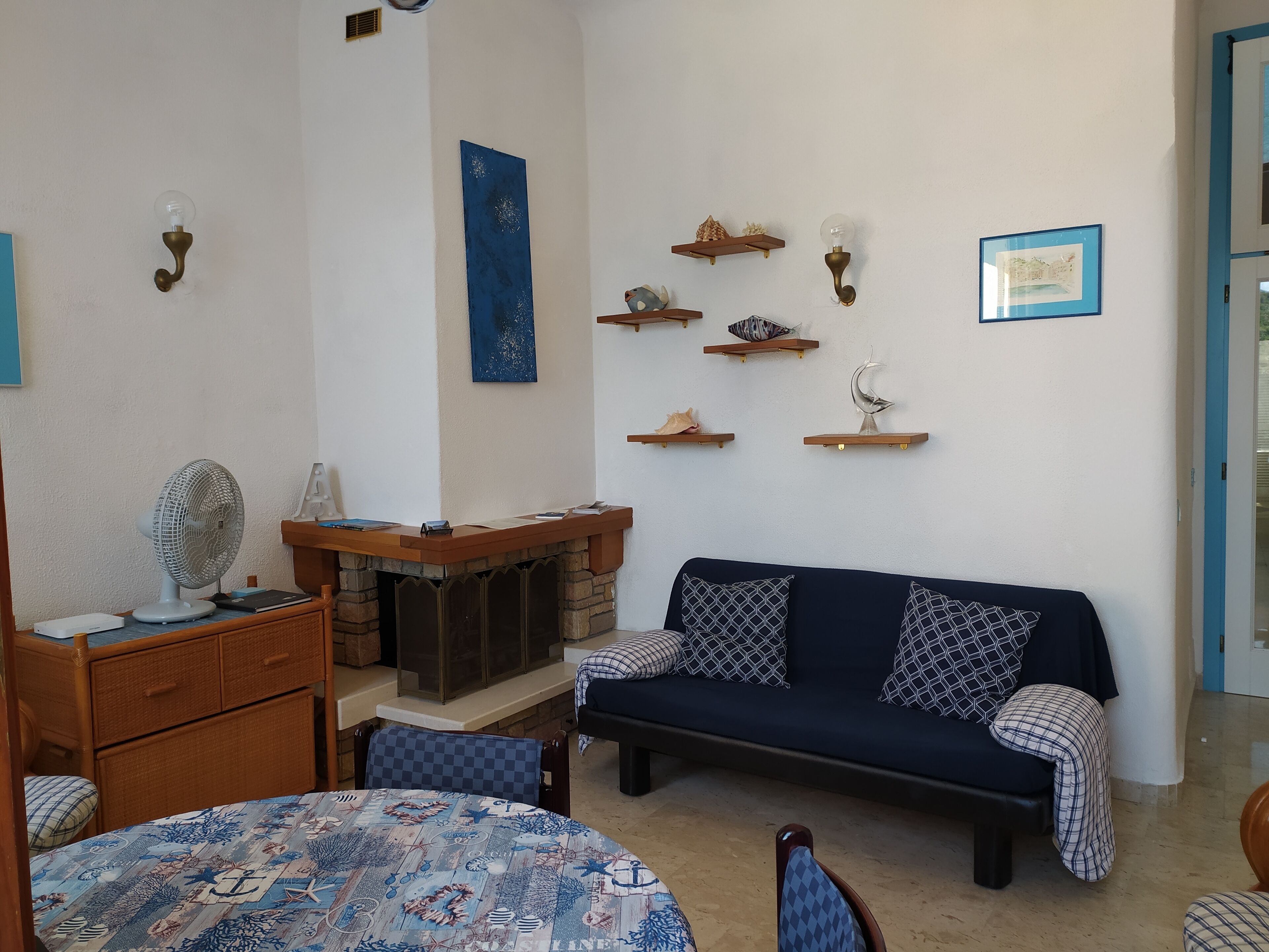 Een gezellige woonkamer met een bank, een tafel en een ventilator. Er zijn wandplanken met decoratieve objecten.