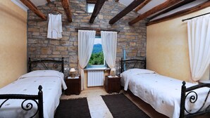 Une chambre rustique aux murs de pierre, avec deux lits, une fenêtre avec des rideaux et une vue sur la verdure.