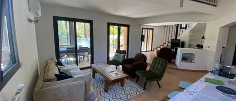A spacious living area with a sofa, armchairs, a coffee table, and a dining table. There are large sliding glass doors leading to an outdoor patio.