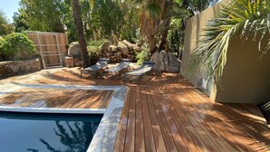 Una terraza de madera con piscina, sillas de descanso y un muro de piedra con plantas.