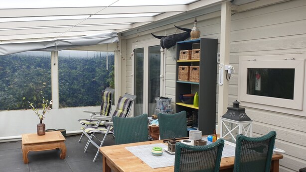 A covered patio with a view of a forested area, wicker furniture, and a bookshelf with baskets. - Family friendly chalet in the Veluwe, Gelderland, Wolfheze (Wolfheze)