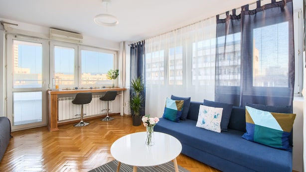 Uma sala de estar moderna com um sofá azul, uma mesa redonda branca e vista para a cidade através de grandes janelas.