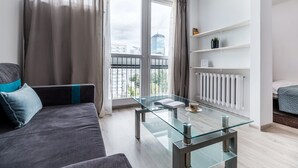 A modern living room with a glass coffee table, grey sofa with teal cushions, a white radiator, and a city view through large windows. - Chlodna 15 Apartment (Warsaw)