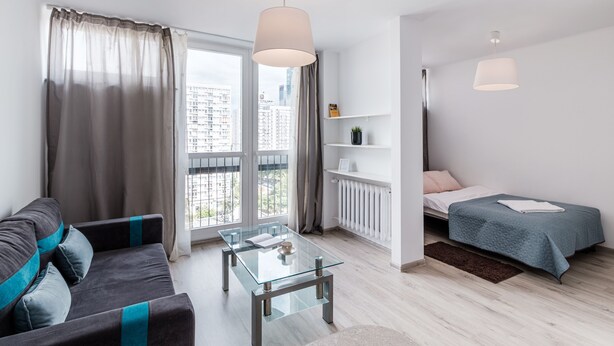 A modern living room with a glass coffee table, a sofa with blue accents, a bed with a blue bedspread, and a view of the city through large windows. - Chlodna 15 Apartment (Warsaw)