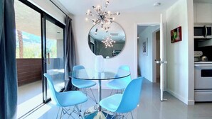 A modern dining area with a glass table and blue chairs, a round mirror on the wall, and a kitchen in the background. - Mod Downtown Escape, Pool, Spa, Tennis, Pickleball (Palm Springs)