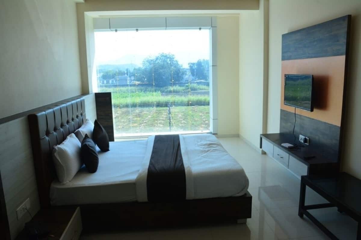 A bedroom with a bed, a TV mounted on the wall, and a window with a view of greenery.