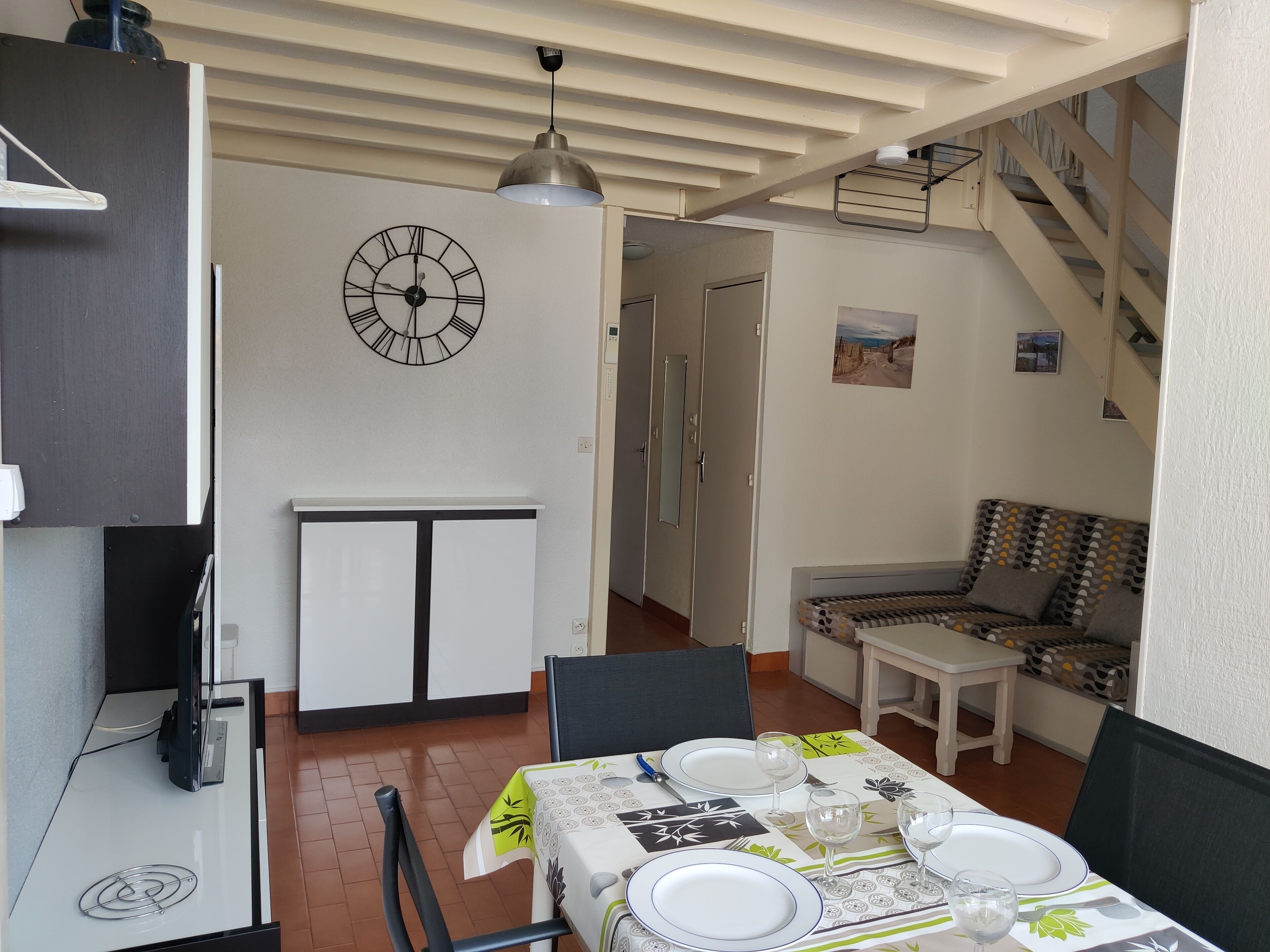 A dining area with a table set for four, a clock on the wall, and a staircase leading to another level.