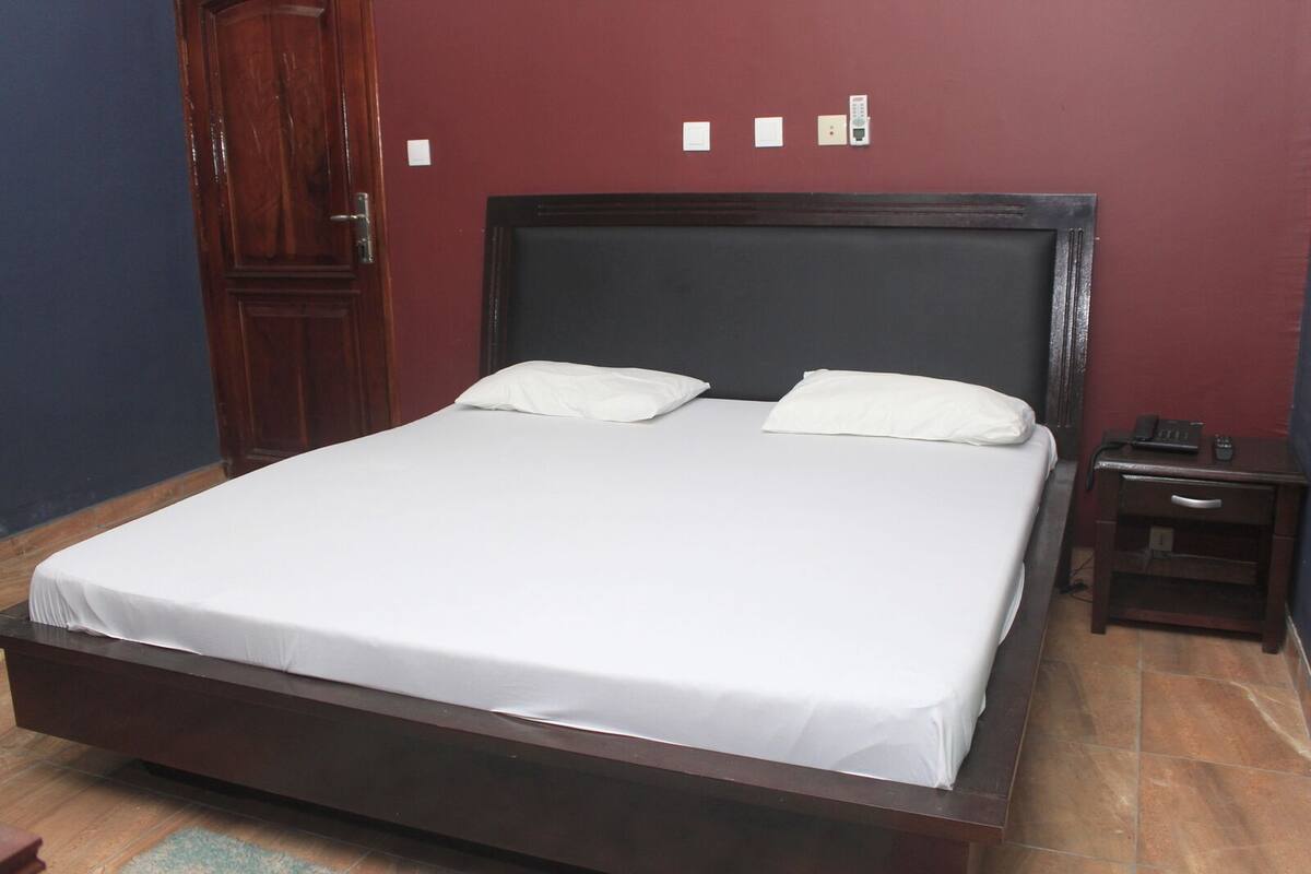 A single bed with a wooden frame and white bedding in a room with a dark wooden door and a small bedside table with a phone and remote control.