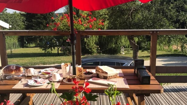 A wooden deck with a picnic setup under a red umbrella, featuring a table with food and drinks, surrounded by red flowers. - Chambre d'Hôte Pivoine (Fralignes)