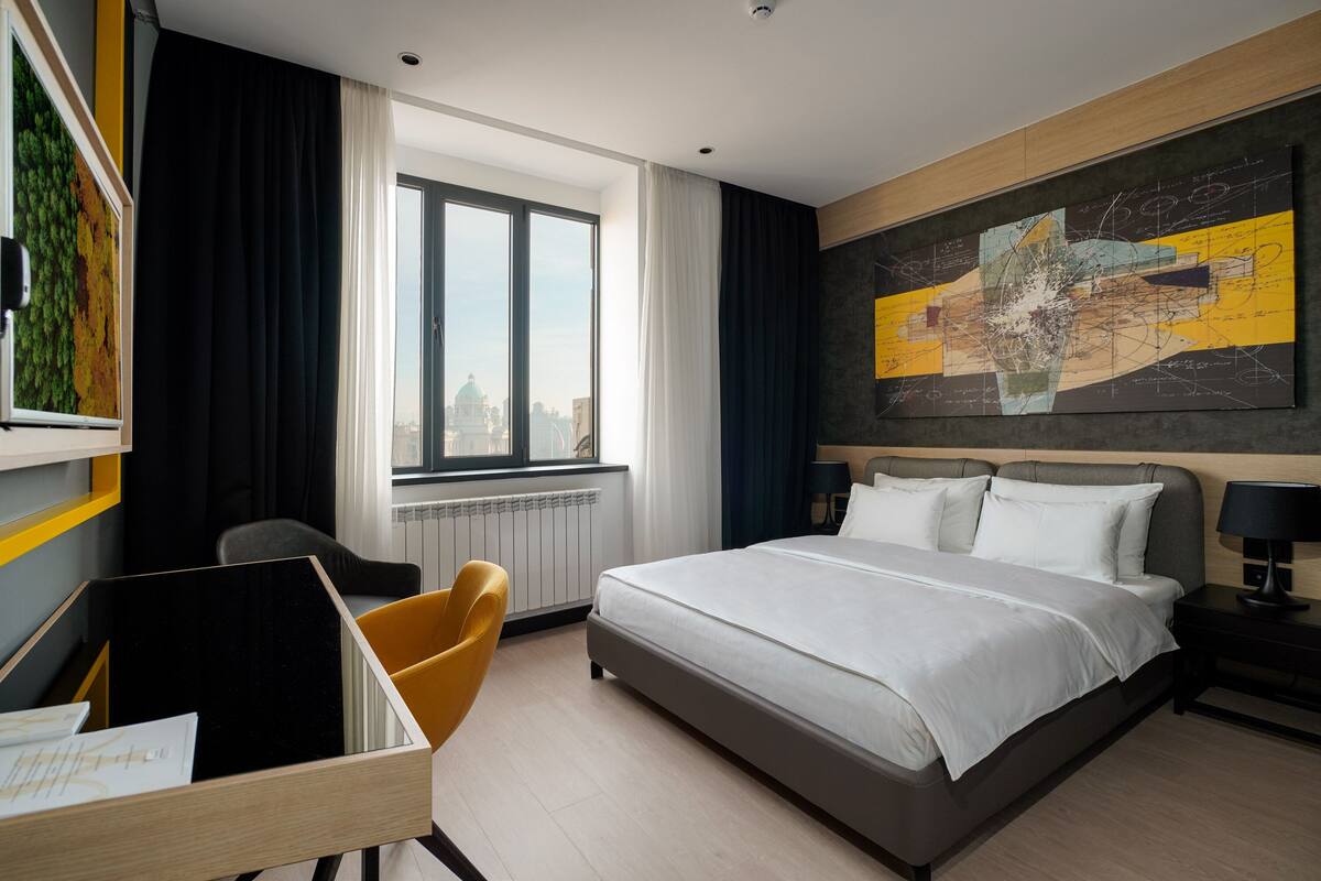 Ein Hotelzimmer mit einem großen Bett, einem Schreibtisch mit Stuhl, einem Fenster mit Vorhängen und Blick auf die Stadt.
