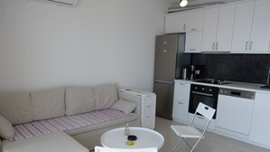 A compact kitchenette with a refrigerator, stove, and dishwasher. There is a small dining area with a table and two chairs. A sofa with a striped cushion is also visible. - Apartment NoStress (Omis)