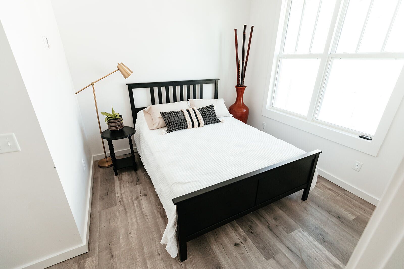 A modern bedroom with a black bed frame, white bedding, a small bedside table with a lamp, a potted plant, and a vase with sticks.