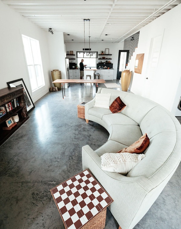 Un vaste espace de vie avec un canapé blanc aux lignes courbes, une table basse à carreaux et une cuisine en arrière-plan.