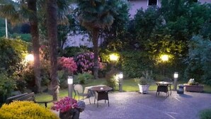 An outdoor patio area with tables and chairs, surrounded by lush greenery and flowering plants. - Hotel Corona (Cuvio)