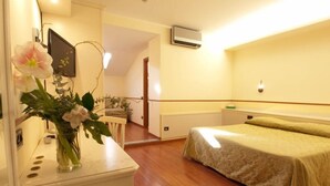 A bedroom with a bed, a small table with a vase of flowers, a wall-mounted air conditioner, and a wooden floor. - Hotel Corona (Cuvio)