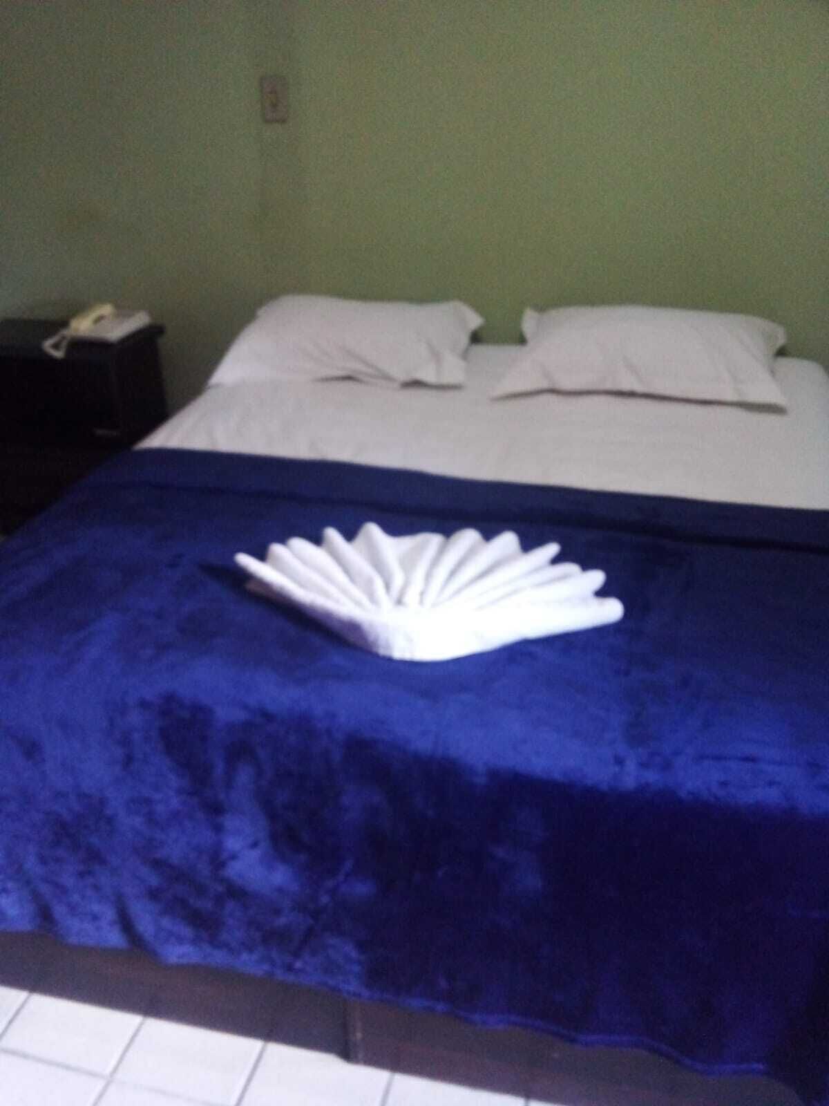 A bed with a blue bedspread and white pillows, a white towel folded on the bed, and a black nightstand with a phone.