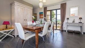 Uma área de refeições com uma mesa de madeira, cadeiras brancas e um armário. Há uma janela com cortinas, um quadro na parede e um abajur sobre uma mesinha.