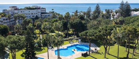A resort with a large pool surrounded by lush greenery and palm trees, overlooking a clear blue sea.