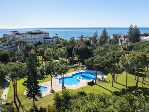 A resort with a large pool surrounded by lush greenery and palm trees, overlooking a clear blue sea.