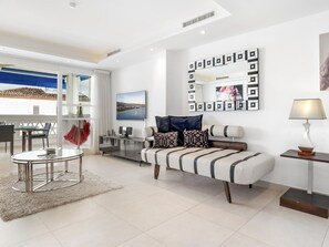 Un salon moderne avec un canapé, une table basse et un téléviseur. Il y a un balcon avec vue sur la mer.