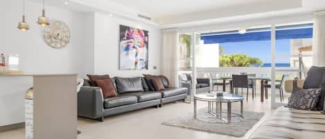 A modern living room with a grey sofa, a coffee table, and a dining area with a view of the sea.