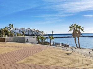 A coastal promenade with a paved walkway, palm trees, and a view of a white building complex by the sea.