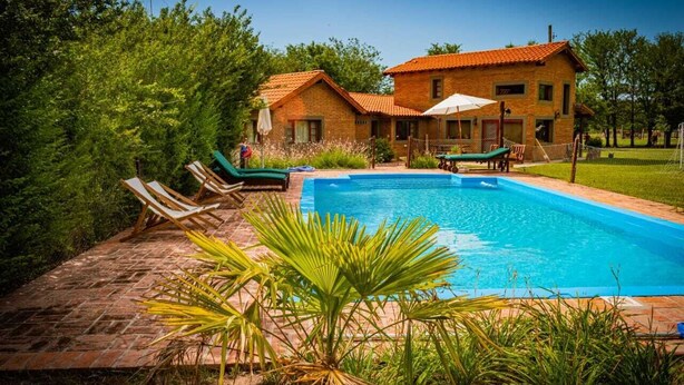 A swimming pool with lounge chairs, a brick pathway, and a house with a tiled roof. - Hotel de campo granja el aromo (Nono)