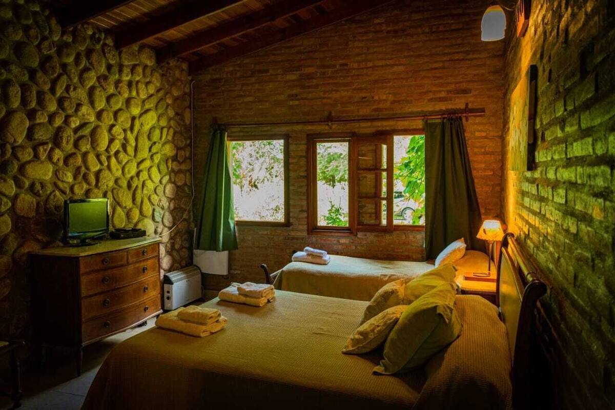 Une chambre rustique avec un mur en pierre, deux lits, une commode en bois, un téléviseur et une fenêtre avec des rideaux.