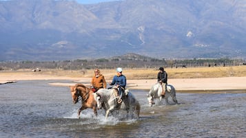 Tres personas cabalgando a caballo por un río, con montañas al fondo.