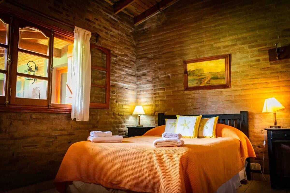 Une chambre rustique avec un mur en briques, un lit avec une literie orange, deux lampes de chevet et un tableau accroché au mur.