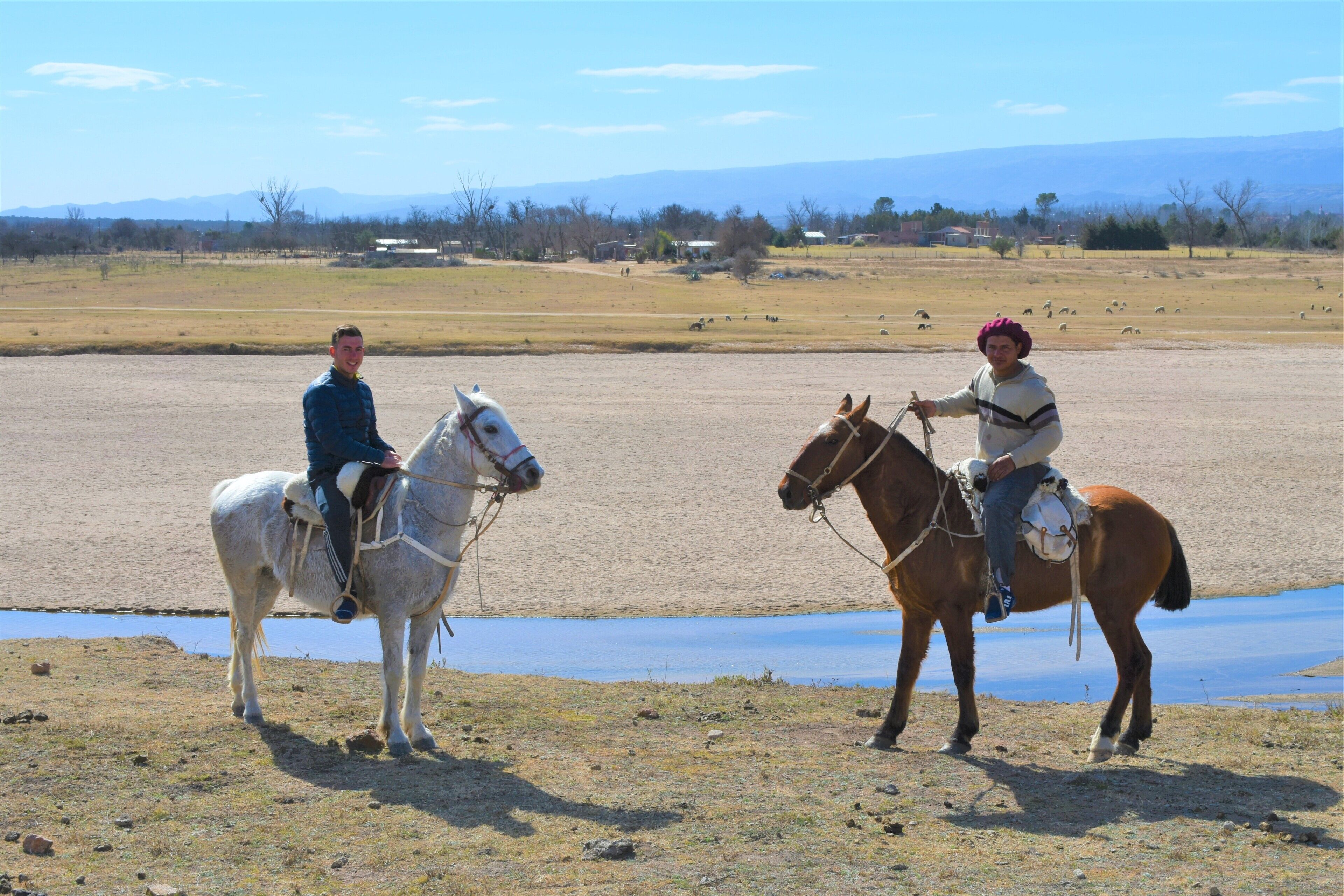 Deux personnes à cheval, évoluant sur un champ aride et dégagé, avec une étendue d’eau et des montagnes lointaines en arrière-plan.