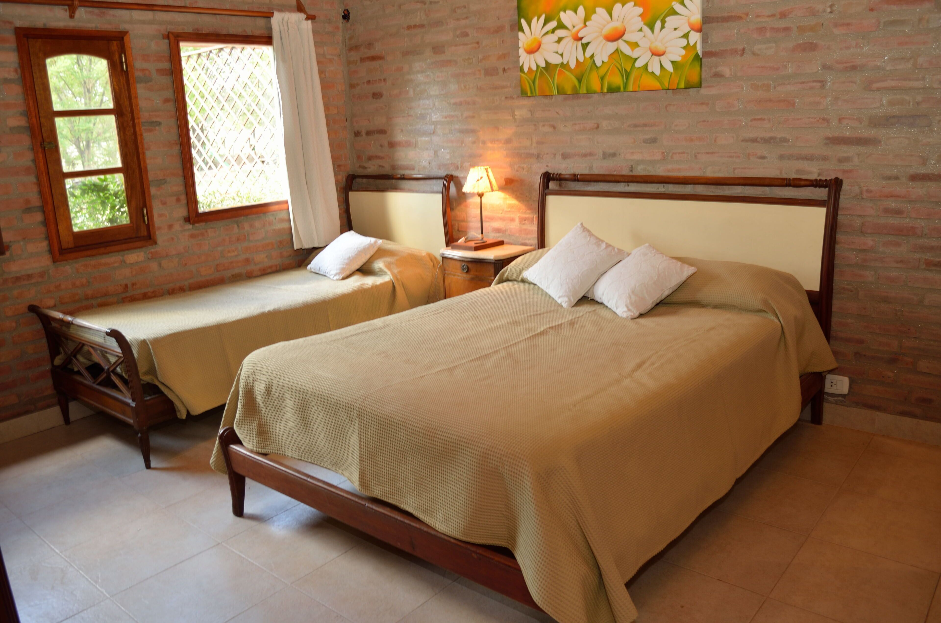 A room with two single beds, a wooden bench, a nightstand with a lamp, and a painting of daisies on the brick wall.