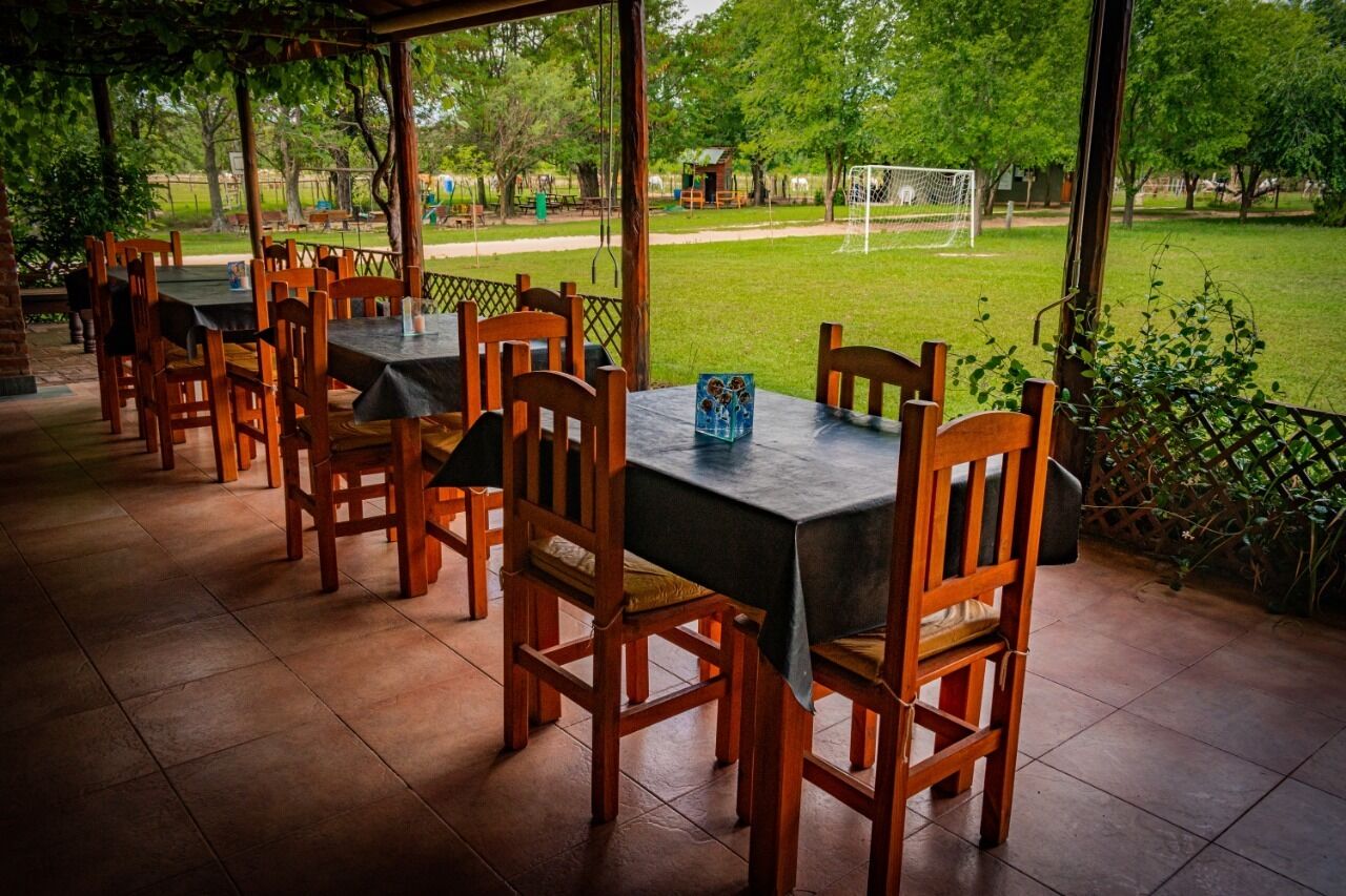 Un patio al aire libre con mesas y sillas de madera, un campo de fútbol al fondo y un paseo techado.