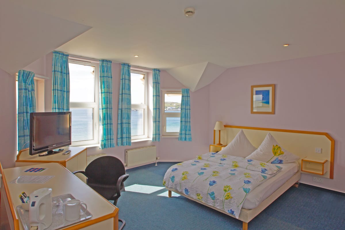 A hotel room with a bed, a desk with a computer and a chair, a TV, and a window with curtains.